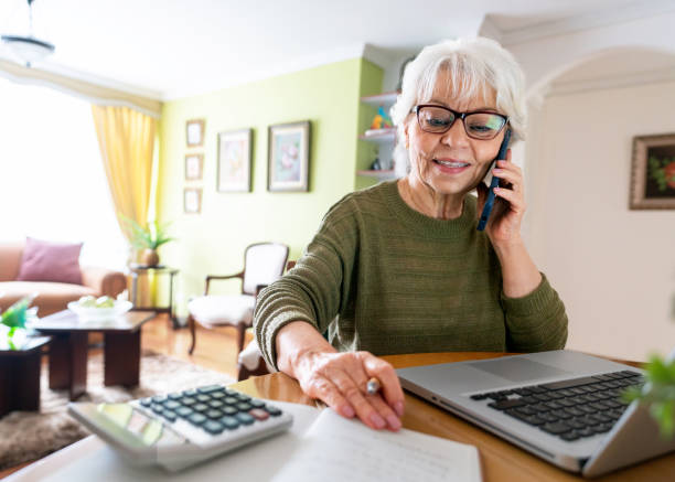 A senior woman talks on the phone while sitting in front of an open laptop and calculator.