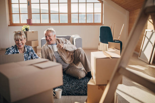 An older couple sitting on the floor packing boxes to move and drinking from coffee mugs.