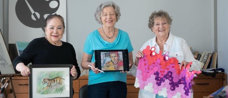 Three senior women in an art studio holding up their own works of art at Moldaw Residences.