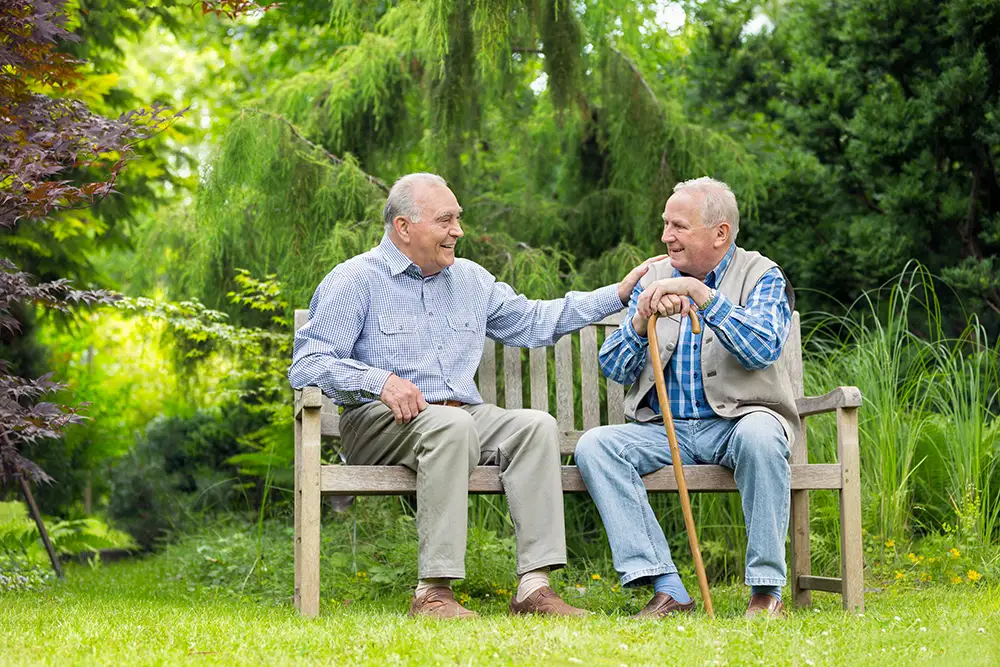 Two senior men sit together on a wooden bench in a garden, smiling and chatting while enjoying outdoor relaxation as Moldaw residents in independent living.